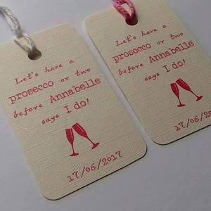 Könnte beinhalten: Zwei weiße Geschenkanhänger mit rosa Kordel. Der Text auf den Anhängern lautet "Let's have a prosecco or two before Annabelle says I do!" mit einem Datum und zwei rosa Sektgläsern.