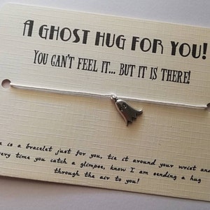 Op de afbeelding: Een wit koordarmband met een zilveren spookbedel. De kaart luidt "Een spook knuffel voor jou! Je kunt het niet voelen... maar het is er!" Hier is een armband speciaal voor jou, bind het om je pols en elke keer dat je een glimp opvangt, weet dan dat ik je een knuffel door de lucht stuur!