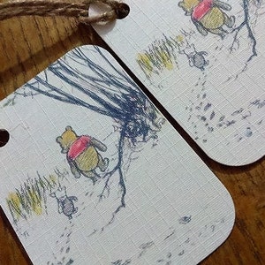 May include: Two Winnie the Pooh gift tags with a white background and brown twine. The tags feature a black and white illustration of Winnie the Pooh walking through the Hundred Acre Wood. The text on the tag reads "Pooh Bear".
