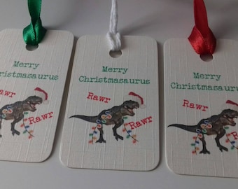 May include: Three rectangular gift tags with a dinosaur wearing a Santa hat and Christmas lights. The tags read "Merry Christmasaurus" and "Rawr" in red and green. Each tag has a different colored ribbon.