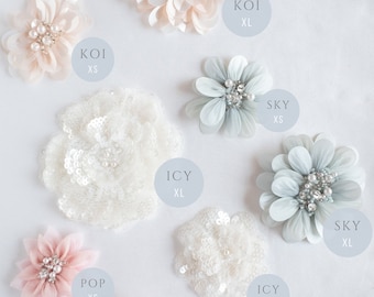 May include: Assortment of eight fabric flower embellishments in various colors and sizes. The flowers are made of delicate fabric and feature pearl accents. The flowers are labeled with names like "Koi", "Sky", "Icy", and "Pop" and sizes like "XS" and "XL".