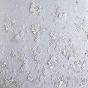 以下が含まれることがあります： 白い生地に、小さな白いビーズとスパンコールの繊細な模様が施されています。生地は透き通るような、ほぼ透明な質感です。