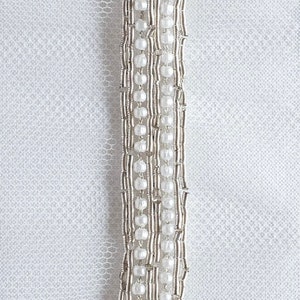 May include: A close-up of a white fabric with a silver beaded trim. The trim is made up of small, round beads that are strung together in a repeating pattern.