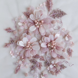 Puede incluir: Un grupo de flores de tela rosa y blanca con adornos de perlas y cristales. Las flores están dispuestas sobre un fondo blanco.