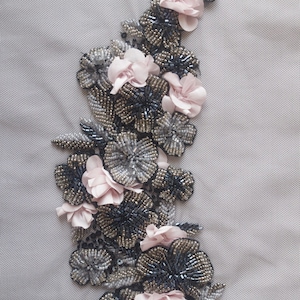 Puede incluir: Un delicado diseño floral con flores de tela rosa y cuentas plateadas y negras. El diseño está bordado sobre una tela gris claro.