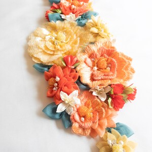 Puede incluir: Un grupo de flores de tela hechas a mano en tonos naranja, amarillo y blanco. Las flores est&aacute;n dispuestas en forma de cascada y presentan delicados detalles de cuentas.