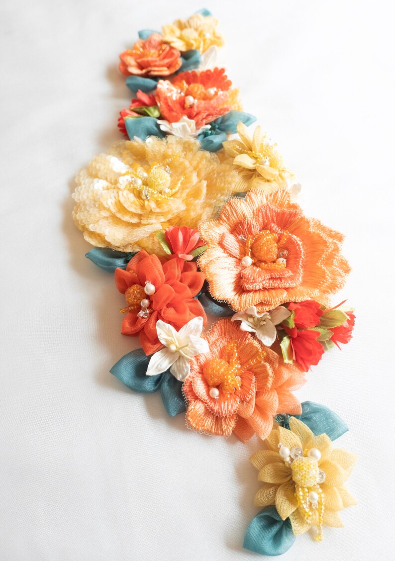 Puede incluir: Un grupo de flores de tela hechas a mano en tonos naranja, amarillo y blanco. Las flores est&aacute;n dispuestas en forma de cascada y presentan delicados detalles de cuentas.
