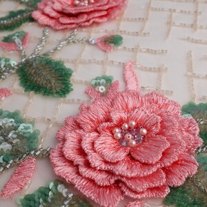 Pu&ograve; includere: Primo piano di un delicato ricamo floreale rosa su uno sfondo di tessuto bianco. Il disegno presenta due rose rosa con foglie verdi e accenti di perle. Il tessuto ha un motivo a griglia di piccole perline bianche.