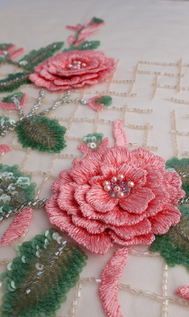 Pu&ograve; includere: Primo piano di un delicato ricamo floreale rosa su uno sfondo di tessuto bianco. Il disegno presenta due rose rosa con foglie verdi e accenti di perle. Il tessuto ha un motivo a griglia di piccole perline bianche.