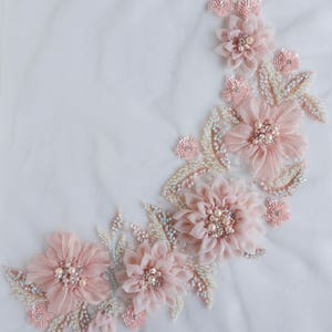 May include: A delicate floral applique with pink fabric flowers and pearl accents. The applique is sewn onto a white mesh fabric.