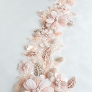 May include: A delicate floral applique with blush pink fabric flowers, pearl beads, and silver sequins. The applique is perfect for adding a touch of elegance to a wedding dress, bridal gown, or other special occasion garment.