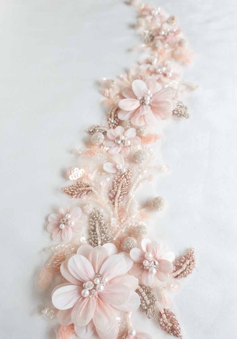 May include: A delicate floral applique with blush pink fabric flowers, pearl beads, and silver sequins. The applique is perfect for adding a touch of elegance to a wedding dress, bridal gown, or other special occasion garment.