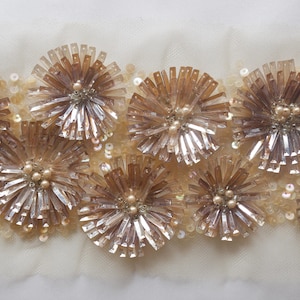 May include: A close-up of a decorative trim with a repeating pattern of gold-toned flower shapes. The flowers are made of small, shiny, metallic pieces that resemble sequins or beads.