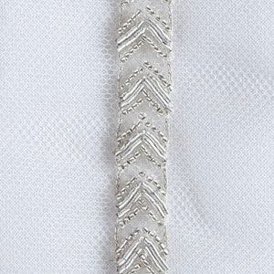 May include: A white fabric with a silver beaded trim. The trim is made of small, silver beads that are arranged in a chevron pattern.
