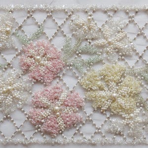 May include: A rectangular piece of fabric with a floral design. The flowers are in pastel colors, including pink, blue, and yellow, and are made of beads. The background is a white, diamond-patterned net. The flowers have pearl centers.