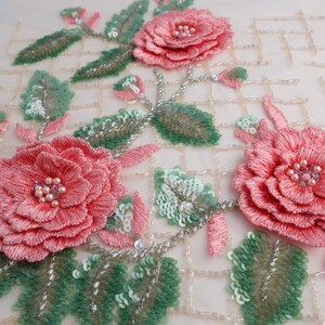 Pu&ograve; includere: Primo piano di un tessuto delicato con rose rosa ricamate e foglie verdi. Il tessuto &egrave; impreziosito da paillettes e perline, creando un design lussuoso e intricato.