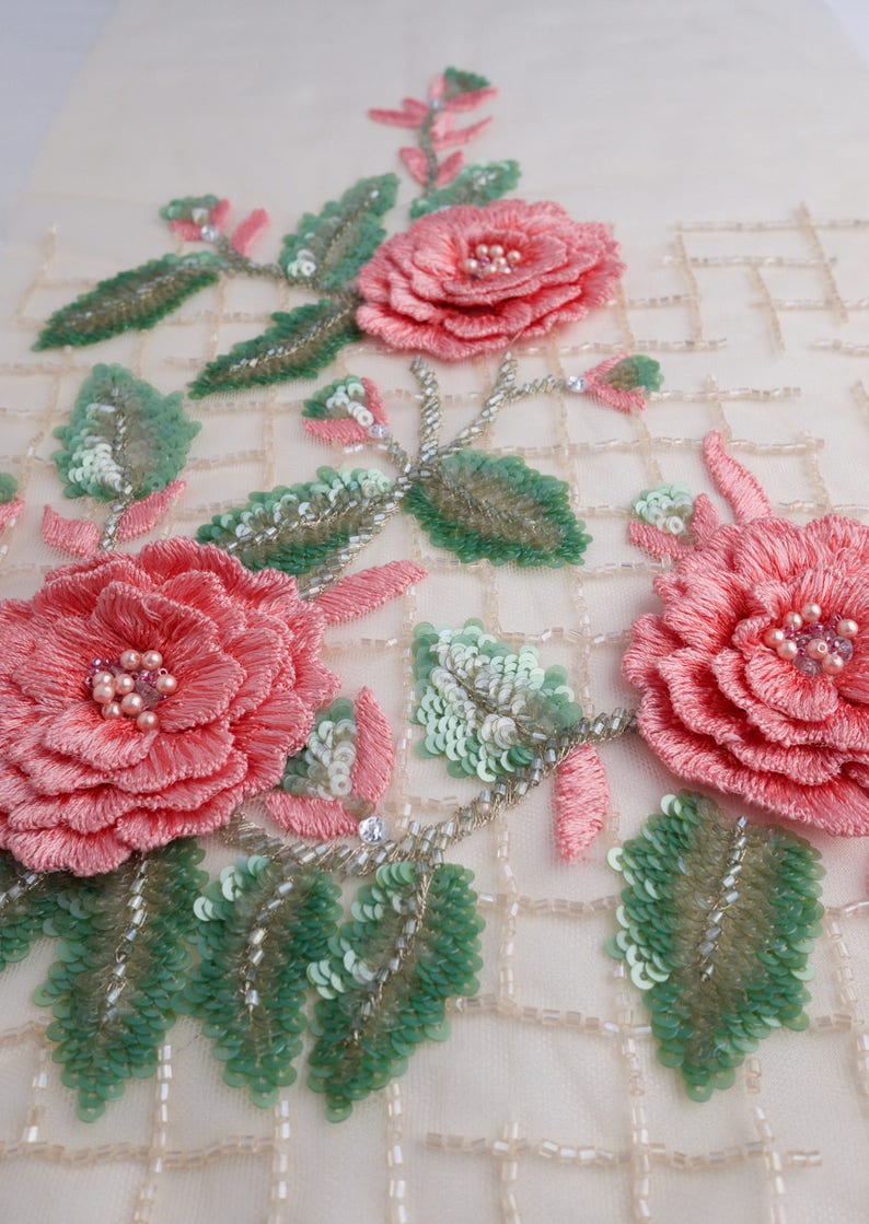 Pu&ograve; includere: Primo piano di un tessuto delicato con rose rosa ricamate e foglie verdi. Il tessuto &egrave; impreziosito da paillettes e perline, creando un design lussuoso e intricato.