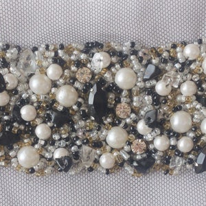 May include: A close-up of a beaded trim with white pearls, black beads, and gold beads. The trim is perfect for adding a touch of elegance to any project.