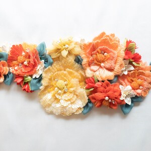 Puede incluir: Un grupo de flores de tela en tonos naranja, amarillo y blanco, con detalles de perlas y detalles de cinta azul.