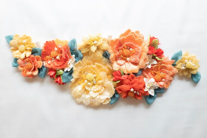 Puede incluir: Un grupo de flores de tela en tonos naranja, amarillo y blanco, con detalles de perlas y detalles de cinta azul.