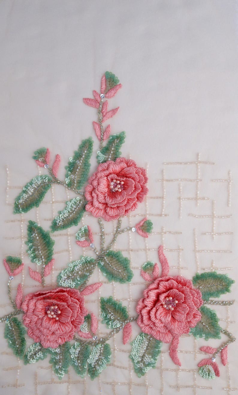 Pu&ograve; includere: Primo piano di delicati fiori ricamati rosa con foglie verdi su uno sfondo di tessuto bianco. I fiori sono realizzati con piccole perline e paillettes.