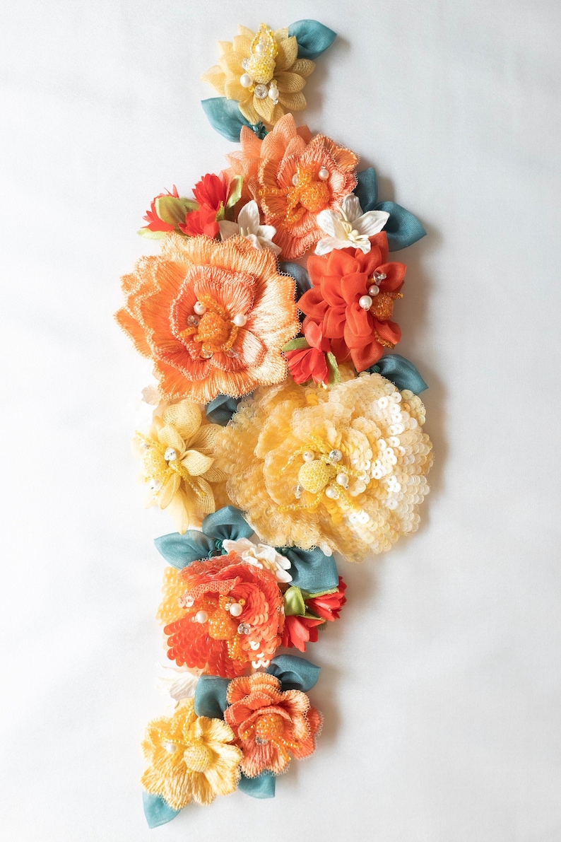 Puede incluir: Un grupo de flores de tela naranja, amarilla y blanca con detalles de perlas y hojas verde turquesa. Las flores est&aacute;n dispuestas en l&iacute;nea vertical y est&aacute;n hechas de diferentes materiales, incluyendo lentejuelas, tul y cinta.