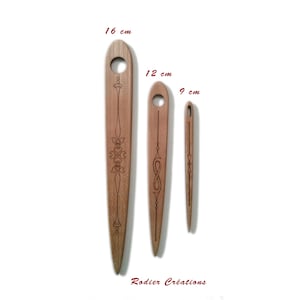 Peut inclure: Trois navettes en bois de différentes tailles, 16 cm, 12 cm et 9 cm de long, avec des motifs gravés. Chaque navette a un trou en haut. Les navettes sont de couleur marron clair. Le texte "Rodier Créations" est en bas.