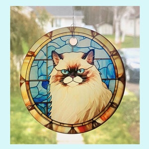 May include: Round stained glass window art featuring a Ragdoll cat with blue eyes and a cream and brown coat. The cat is set against a blue and gold stained glass background. A small metal loop is attached for hanging.
