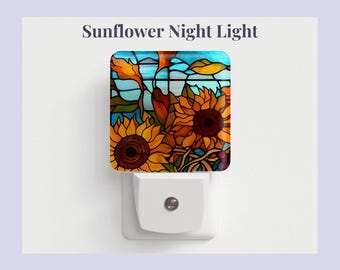 Pode incluir: Uma luz noturna com um design de vitral que apresenta girassóis em tons de amarelo, laranja e marrom. A luz noturna é branca e tem um design plug-in.