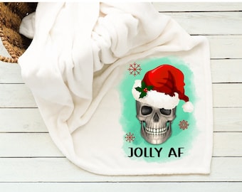 Pode incluir: Cobertor de lã branca com uma caveira usando um chapéu de Papai Noel sobre um fundo turquesa. O texto "Jolly AF" está impresso abaixo da caveira.