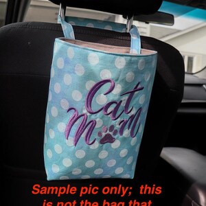 K&ouml;nnte beinhalten: Eine blaue und wei&szlig;e gepunktete Stofftasche mit dem gestickten Text "Cat Mom" in Lila. Die Tasche h&auml;ngt an der Kopfst&uuml;tze eines Autositzes.