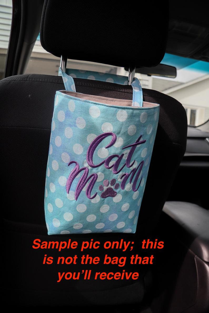 K&ouml;nnte beinhalten: Eine blaue und wei&szlig;e gepunktete Stofftasche mit dem gestickten Text "Cat Mom" in Lila. Die Tasche h&auml;ngt an der Kopfst&uuml;tze eines Autositzes.