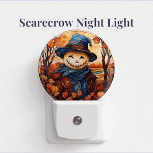Peut inclure: Une veilleuse blanche avec une image ronde et colorée d'un épouvantail avec un visage de citrouille, portant un chapeau et une écharpe bleus, entouré de citrouilles et de feuilles d'automne. Le texte "Scarecrow Night Light" est au-dessus de l'image.
