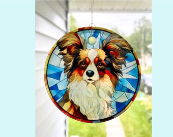 Pode incluir: Um apanhador de sol de vitral com um cão papillon de pêlo castanho, branco e laranja. O rosto do cão é visível e o fundo é um desenho de vitral azul e branco.