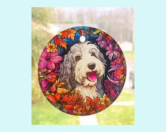 May include: A round glass ornament featuring a colorful illustration of a bearded collie dog surrounded by autumn leaves and flowers. The dog has a white and gray coat and is smiling with its tongue sticking out.