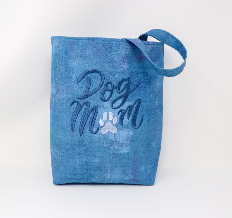 K&ouml;nnte beinhalten: Blaue Tragetasche mit dem gestickten Text "Dog Mom" in Wei&szlig; und Blau mit einem wei&szlig;en Pfotenabdruck.