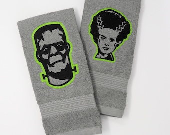 Pode incluir: Duas toalhas de cozinha cinzas com imagens bordadas do monstro de Frankenstein e sua noiva. O bordado é preto com um contorno verde brilhante.
