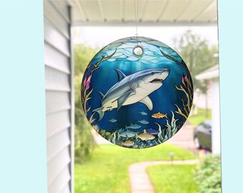 以下が含まれることがあります： 青い背景に白いサメが海を泳いでいる様子が描かれた丸いガラスのオーナメント。サメの周りはサンゴや他の魚で囲まれている。オーナメントには銀色のハンガーが付いている。