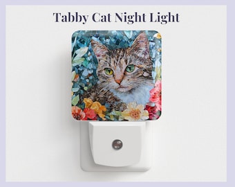 Pode incluir: Uma luz noturna com um gato tigrado de olhos verdes rodeado de flores coloridas. A luz noturna é branca e tem uma tomada padrão.