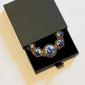 May include: A gold necklace with blue and white porcelain pendants, depicting windmills, is displayed in an open black box. The necklace features a delicate, filigree design around each pendant, offering an elegant appearance.