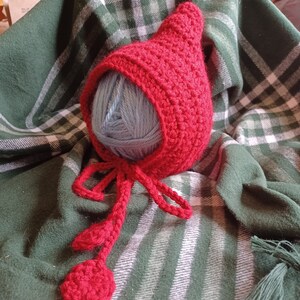 May include: Red crocheted baby bonnet with a long tie and two red pom-poms. The bonnet is lying on a green and white plaid blanket.
