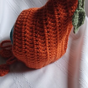 May include: A handmade orange crochet pumpkin hat with a green stem and leaves. The hat is made of textured yarn and has a drawstring. The hat is likely for a baby or toddler.