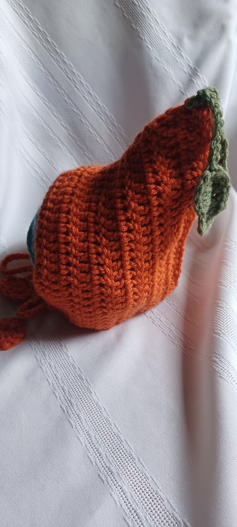 May include: A handmade orange crochet pumpkin hat with a green stem and leaves. The hat is made of textured yarn and has a drawstring. The hat is likely for a baby or toddler.