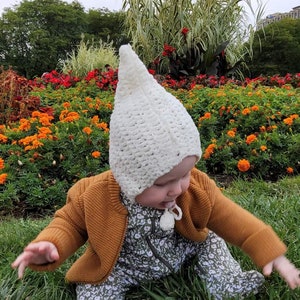 May include: A baby wearing a white crocheted hat, a mustard-colored cardigan, and a floral print jumpsuit, sitting in the grass. The background features a vibrant flower bed with orange and red blooms, and tall green plants.