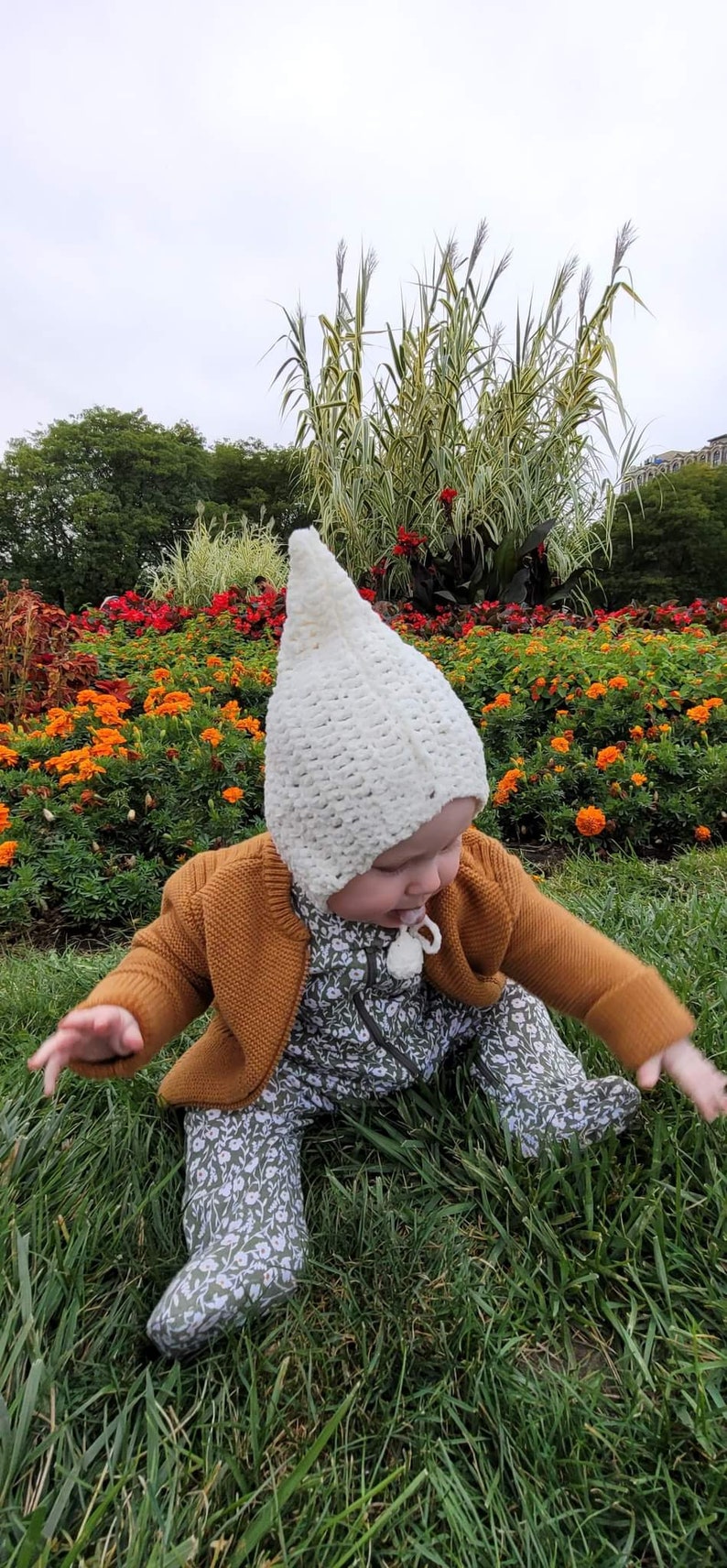 May include: A baby wearing a white crocheted hat, a mustard-colored cardigan, and a floral print jumpsuit, sitting in the grass. The background features a vibrant flower bed with orange and red blooms, and tall green plants.