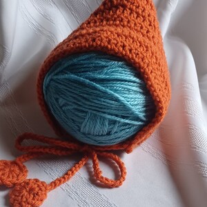 May include: An orange crocheted baby bonnet with a green stem detail, covering a ball of teal yarn. The bonnet has a long orange tie with two crocheted circles at the ends. The item is on a white, textured surface.