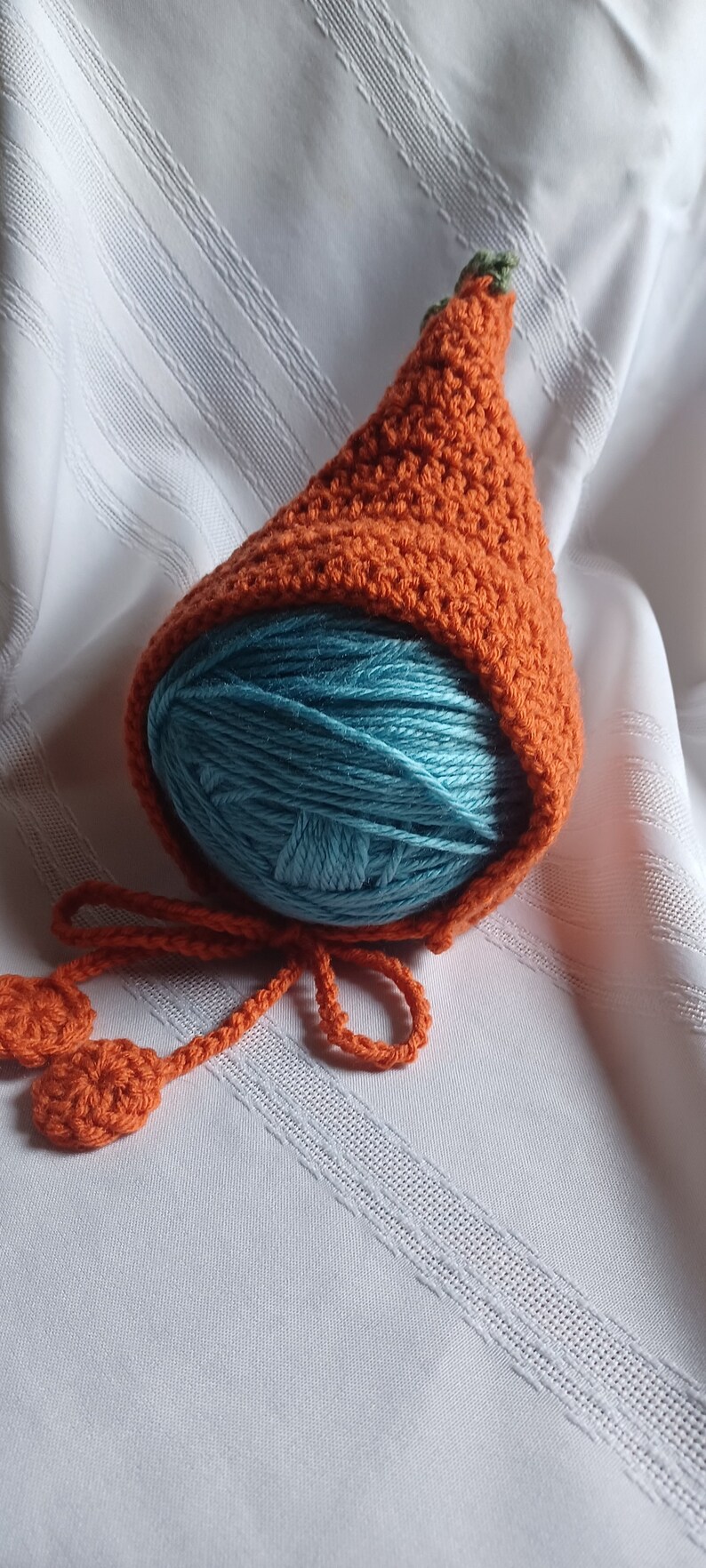 May include: An orange crocheted baby bonnet with a green stem detail, covering a ball of teal yarn. The bonnet has a long orange tie with two crocheted circles at the ends. The item is on a white, textured surface.