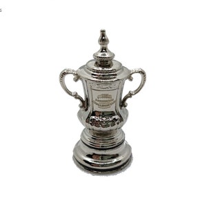 Könnte beinhalten: Eine silberne Nachbildung des FA Cup-Pokals. Der Pokal hat zwei Henkel und einen runden Sockel. Die Inschrift "The Football Association Challenge Cup" ist an der Seite des Pokals sichtbar.
