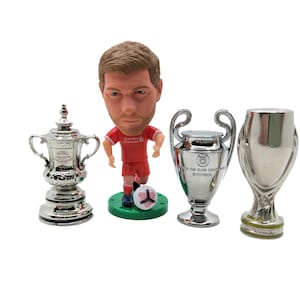 May include: A silver miniature replica of the FA Cup, a silver miniature replica of the UEFA Champions League trophy, and a silver miniature replica of the UEFA Europa League trophy, along with a figurine of a soccer player wearing a red jersey.