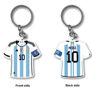 Op de afbeelding: Een sleutelhanger in de vorm van een wit en lichtblauw voetbaltenue met nummer 10 op de voor- en achterkant. De achterkant van het tenue heeft "Messi" boven het nummer. De sleutelhanger is gemaakt van metaal en heeft een zilveren afwerking.